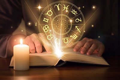 Hands placed on an open book with zodiac signs in a circle and stars floating above.